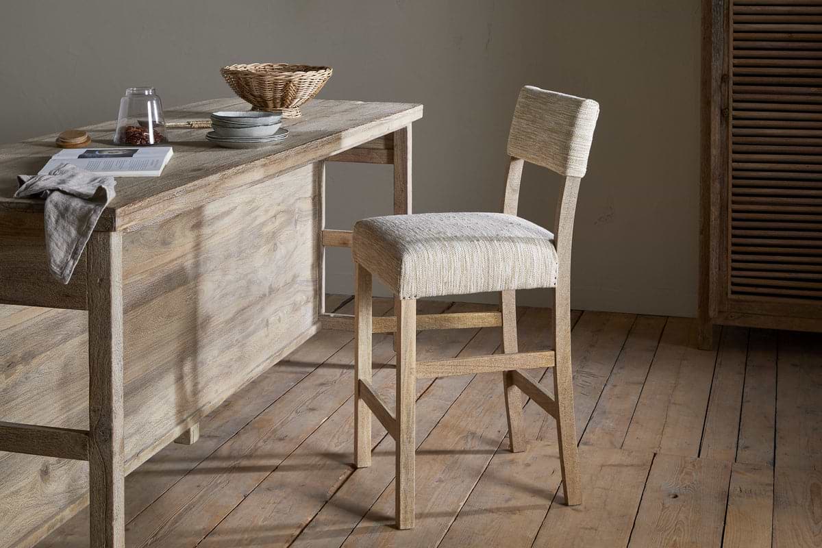 A rustic kitchen features the nkuku Zoha Cotton & Jute Upholstered Counter Chair in natural, set at a wooden table styled with a towel, glass, pitcher, book, plates, and wicker basket amid neutral minimalist décor.