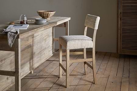 A rustic kitchen features the nkuku Zoha Cotton & Jute Upholstered Counter Chair in natural, set at a wooden table styled with a towel, glass, pitcher, book, plates, and wicker basket amid neutral minimalist décor.