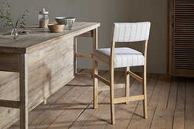 A rustic wooden table with light gray dishes and a vase of green stems sits on a wooden floor. Beside the table is the nkuku Nidhi Linen Upholstered Counter Chair in Natural, crafted from mango wood with light, striped linen upholstery.