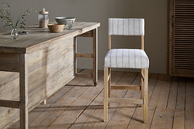The Nidhi Linen Upholstered Counter Chair by nkuku features striped white linen on a mango wood frame. It sits on a wooden floor beside a rustic table topped with greenery, baskets, and a lidded jar in a softly lit, neutral-toned room.