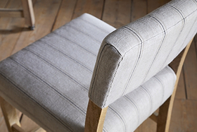 A close-up, angled view of the nkuku Nidhi Linen Upholstered Counter Chair - Natural, featuring light gray linen with thin vertical stripes and a mango wood frame, set on a light brown wooden floor. Another chair appears slightly out of focus in the background.