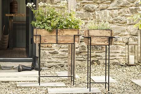 Two rustic reclaimed wooden planter boxes with green and white plants rest on tall nkuku Amazzi Indoor Planter Stands in Natural & Black, placed on a pebble patio before a stone wall. A door, black boot, and lantern enhance the cozy outdoor scene.