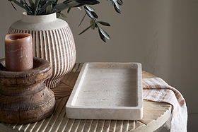 A beige ceramic vase with green leaves, a brown pillar candle on a rustic wooden stand, and the nkuku Ganda Travertine Vanity Tray in cream sit atop a round table with a beige-striped cloth, creating a softly lit, minimalistic ambiance.