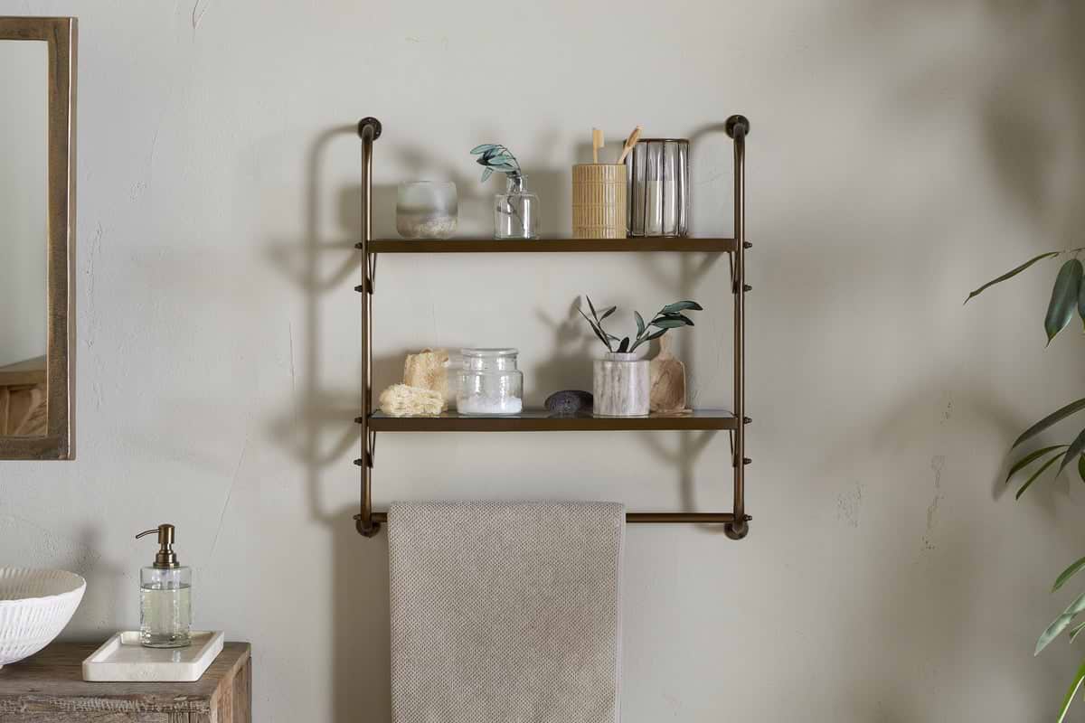 The Karura Wall Shelves with Towel Rail in Antique Brass by nkuku hold jars, a toothbrush holder, cups, a loofah, stones, and small plant cuttings. A beige towel hangs below; to the left sit a mirror and soap dispenser on a wooden counter.