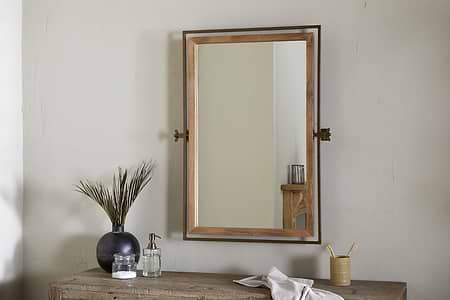 The nkuku Mar Swivel Wall Mirror in Antique Brass & Natural is mounted on a light beige wall above a rustic wooden table, which holds a black vase with green stems, clear soap dispenser, bamboo toothbrush cup, and folded white cloth.