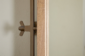 A close-up of the nkuku Mar Swivel Wall Mirror - Antique Brass & Natural shows its brown metal bracket with a wingnut attached, mounted on a light wall. The antique brass edge and visible wood grain highlight its elegant design.