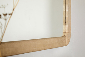 A close-up of the nkuku Vivan Rectangular Grooved Wood Mirror in natural, featuring a hand-carved mango wood frame with textured grooves, mounted on a white wall. Soft-focus dried stems appear on the left.