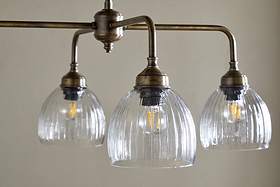 The Kakira Chandelier Pendant by nkuku features antique brass and clear ribbed bell-shaped glass shades. Three exposed bulbs hang from a horizontal rod, creating a vintage-industrial focal point for any space.