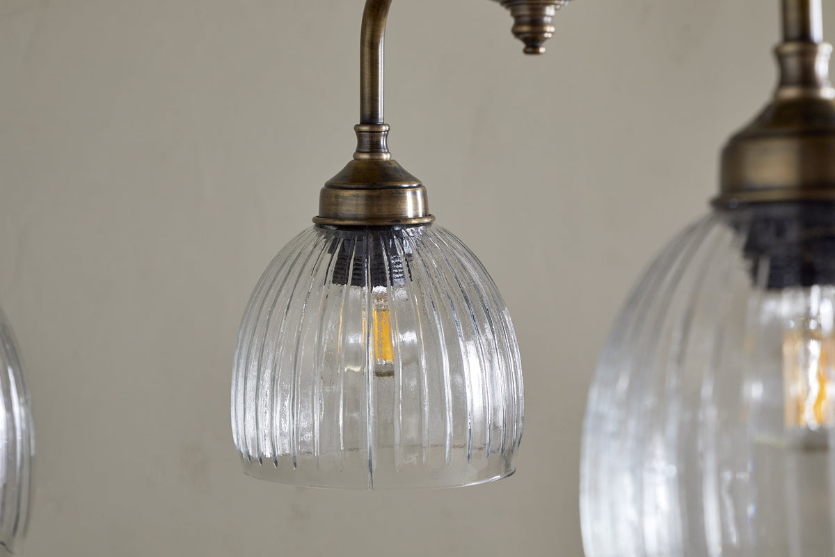 Close-up of the Kakira Chandelier Pendant by nkuku, featuring a clear ribbed glass shade and antique brass fixture. The glowing bulb and neutral background enhance the elegant details of this refined light.