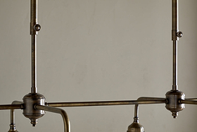 Close-up of the nkuku Kakira Chandelier Pendant in Antique Brass & Clear, featuring visible rods and joints, clean lines, metal hardware, and a muted finish—all set against a plain beige wall for an industrial yet elegant look.