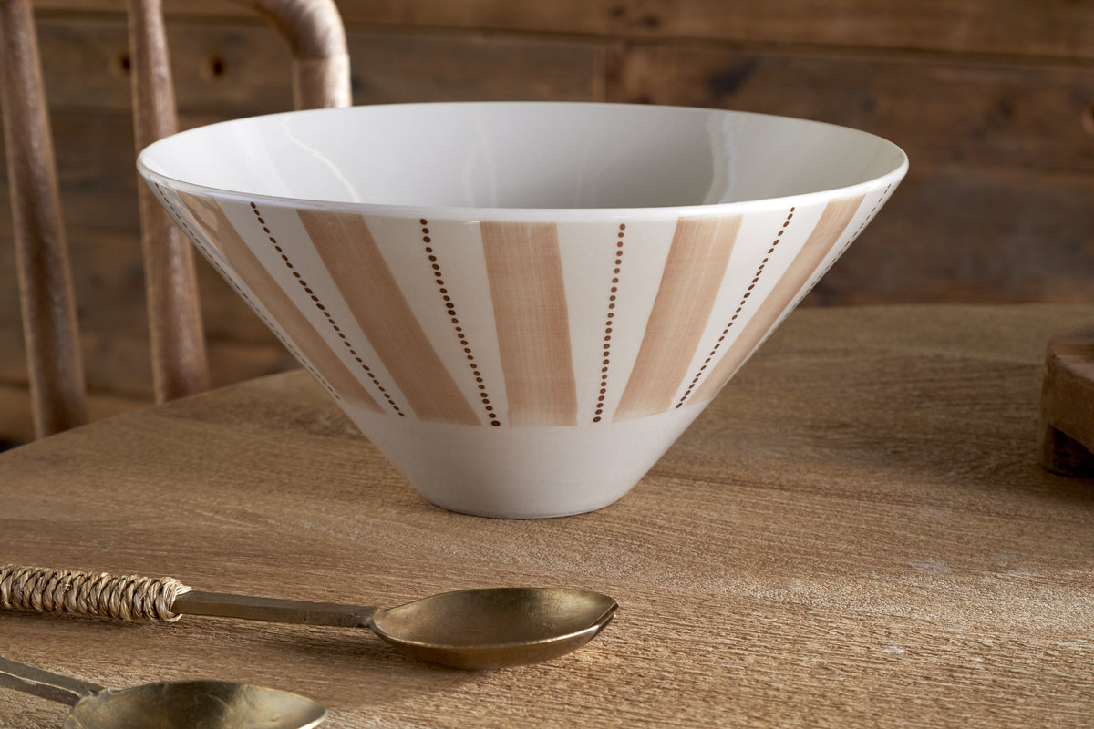 The Patthar Serving Bowl in Cream & Clay (Medium) by nkuku, featuring hand-painted tan vertical stripes, rests on a wooden table beside two brass-colored spoons with woven handles. A wooden chair and paneled wall complete the rustic scene.