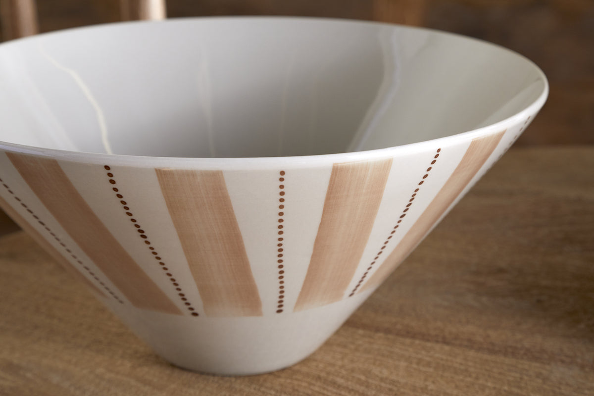 The Patthar Serving Bowl - Cream & Clay - Medium by nkuku sits on a wooden surface. Part of a dinnerware collection, it features vertical beige stripes with hand-painted brown dots, creating a simple geometric pattern.