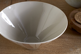 The nkuku Patthar Serving Bowl - Cream & Clay (Medium) features a glossy, light gray ceramic finish and rests on a wooden surface, with part of a woven coaster and another round item visible in this elegant dinnerware collection.