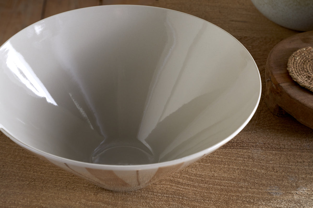 The nkuku Patthar Serving Bowl - Cream & Clay (Medium) features a glossy, light gray ceramic finish and rests on a wooden surface, with part of a woven coaster and another round item visible in this elegant dinnerware collection.