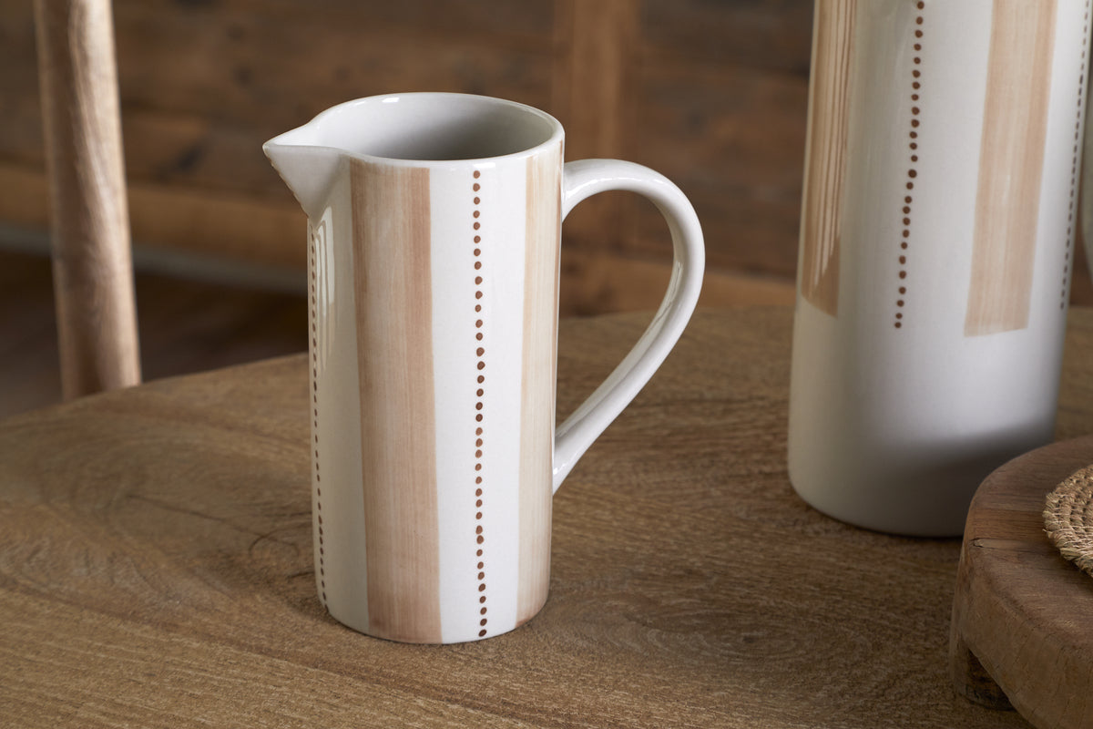 The nkuku Patthar Jug - Cream & Clay - Small, hand-painted with vertical beige stripes and dotted lines, sits on a wooden table alongside pieces from the matching tableware collection and a woven placemat.