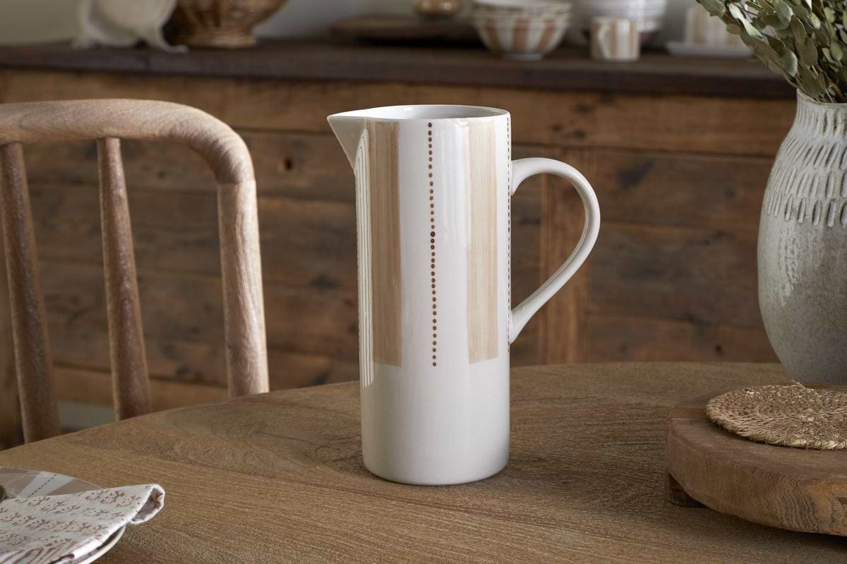 The nkuku Patthar Jug - Cream & Clay - Large, a tall hand-painted ceramic piece with delicate vertical dotted lines, sits on a wooden table set with a napkin, woven placemat, and large vase. A rustic chair and sideboard complete the cozy scene.
