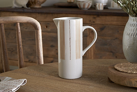 The nkuku Patthar Jug - Cream & Clay - Large, a tall hand-painted ceramic piece with delicate vertical dotted lines, sits on a wooden table set with a napkin, woven placemat, and large vase. A rustic chair and sideboard complete the cozy scene.