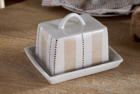 The nkuku Patthar Butter Dish - Cream & Clay is a hand-painted ceramic piece with a looped handle, featuring vertical beige stripes and dotted brown lines. It sits on a wooden table, with kitchen storage plates stacked in the background.