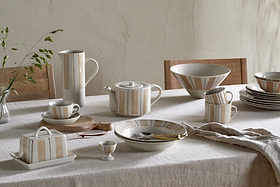 A set table with a neutral cloth displays striped ceramic dinnerware and the hand-painted nkuku Patthar Butter Dish in Cream & Clay. Cutlery, an egg cup, linen napkin, and greenery in a glass vase add to this elegant, casual arrangement.