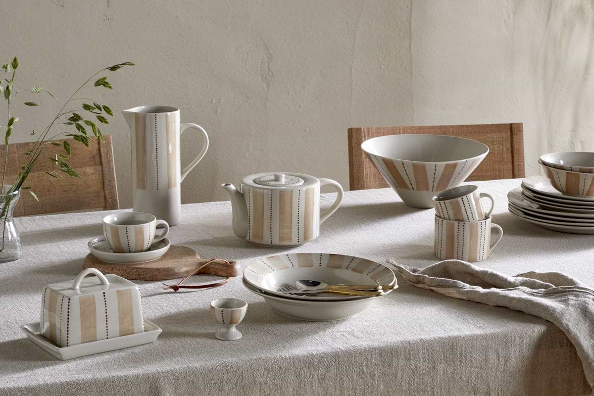 A beige tablecloth covers a table set with nkuku's Patthar Cup & Saucer - Cream & Clay, handmade dinnerware, cutlery, and bowls. Wooden chairs and green foliage bring a natural touch against the textured wall.