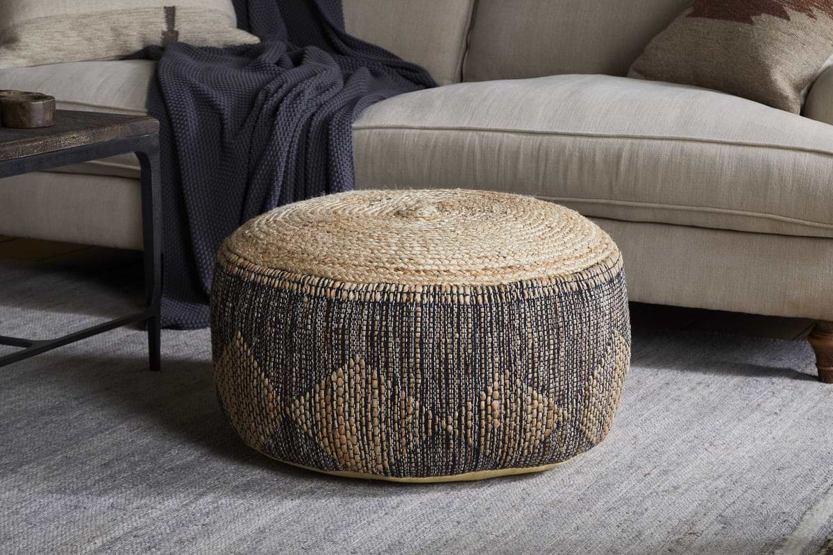 The Kapil Jute Round Pouf - Charcoal by nkuku, featuring a geometric braided design, sits on a light gray rug in front of a beige sofa with a dark blanket, while a wooden coffee table adds to the chic setting.