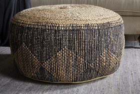 The Kapil Jute Round Pouf - Charcoal by nkuku, featuring a natural tan top and black diamond braided design, sits on a gray rug in front of a beige sofa, adding cozy texture and style to your living space.