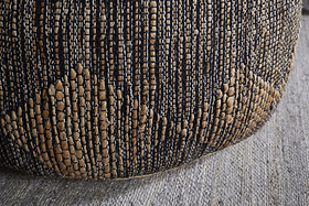 Close-up of the Kapil Jute Round Pouf - Charcoal by nkuku, featuring a braided tan and dark blue jute design with diamond patterns, resting on a textured light gray rug.