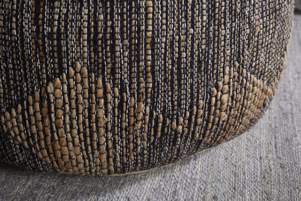 Close-up of the Kapil Jute Round Pouf - Charcoal by nkuku, featuring a braided tan and dark blue jute design with diamond patterns, resting on a textured light gray rug.