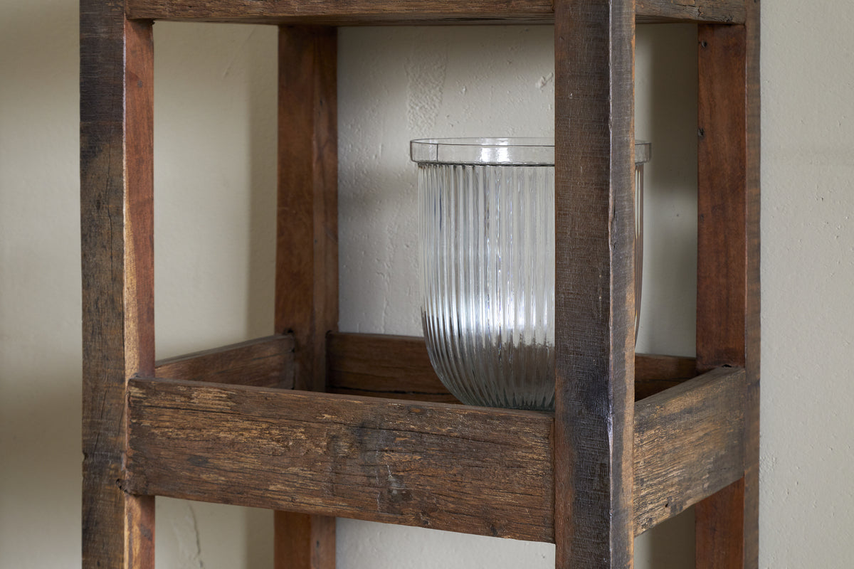 A clear, ribbed glass vase sits on the Akara Reclaimed Wood Storage Caddy - Natural by nkuku, its rustic unfinished look and visible wood grain making it a charming organizer against the plain, light-colored wall.