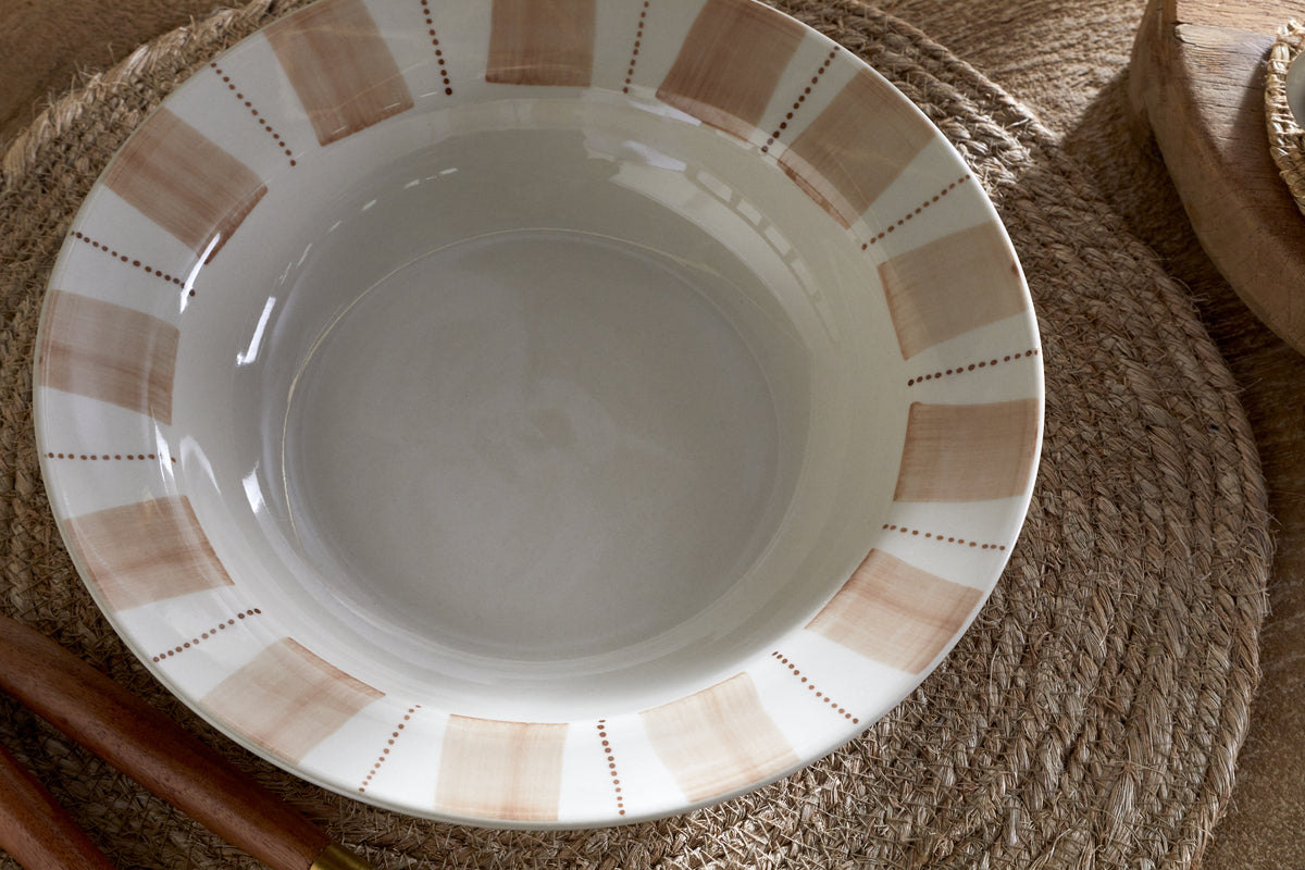 The nkuku Patthar Pasta Bowl in Cream & Clay features tan, brushstroke-style stripes and dotted details on white ceramic, displayed on a woven mat with wooden utensils—an artisanal touch for any dinnerware collection.
