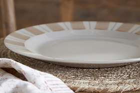 The nkuku Patthar Dinner Plate - Cream & Clay, featuring a shallow round design with a beige and white striped rim, rests on a woven placemat with a patterned cloth nearby and a blurred wooden background.