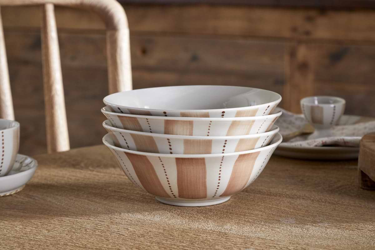 The Patthar Dinnerware Set - Cream & Clay by nkuku features 12 hand-painted ceramic pieces, stacked on a wooden table with rustic surfaces in the background, evoking a cozy farmhouse kitchen vibe.
