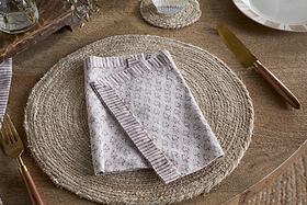 A beige woven placemat sits on a wooden table with an nkuku Peplu Block Print Napkin - Terracotta folded at the center. A gold fork is on the left, a gold knife with a wooden handle on the right, and part of a white plate is visible in the top right corner.