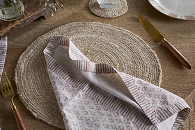 A rustic table setting with a round woven placemat, nkuku Peplu Block Print Napkin in Terracotta, gold cutlery with wooden handles, and a clear glass on a matching coaster atop a wooden tabletop.