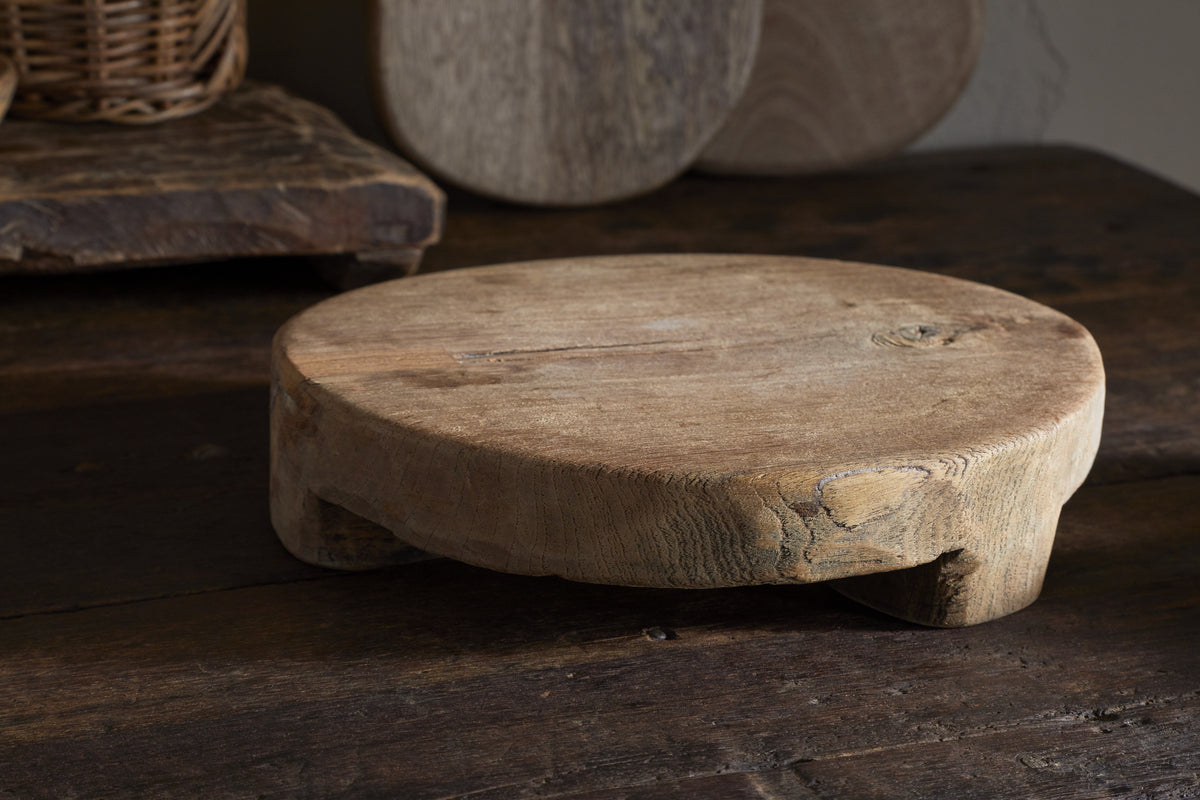 The Kaveeta Board - Natural by nkuku features a round, rustic wood design with visible grain and timeworn details, resting on a dark surface. Reclaimed boards and baskets in the background create an earthy, vintage feel.
