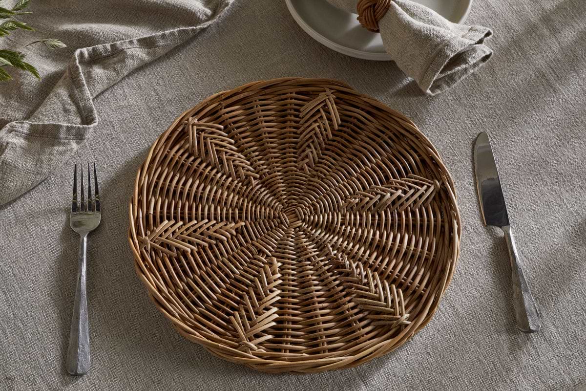 The nkuku Bersama Rattan Placemat - Natural sits on a beige linen tablecloth, with cutlery on each side. A beige napkin in a rattan ring tops stacked plates, creating a rustic dining setting.