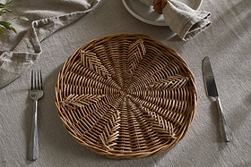 The nkuku Bersama Rattan Placemat - Natural sits on a beige linen tablecloth, with cutlery on each side. A beige napkin in a rattan ring tops stacked plates, creating a rustic dining setting.