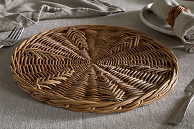 The Bersama Rattan Placemat by nkuku, featuring intricate handwoven details, rests on a light gray tablecloth beside a plate with a napkin in a wicker ring, fork, and knife—creating a rustic dining setting in soft natural light.