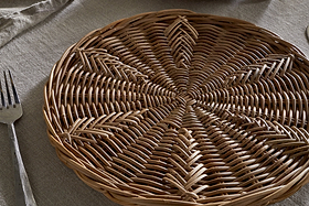 The nkuku Bersama Rattan Placemat - Natural, featuring intricate handwoven details, rests on a beige fabric tablecloth. A metal fork lies to the left, with a folded napkin partly visible in the upper left corner of the table.