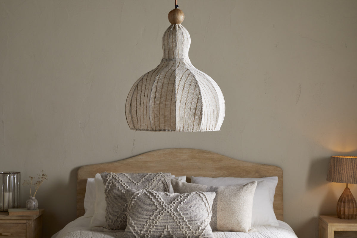 A neutral-toned bedroom features a wooden bed with cream and gray pillows. The nkuku Kela Fabric Pendant in Off White hangs above, while a lamp and glass vase rest on the light wood nightstand.