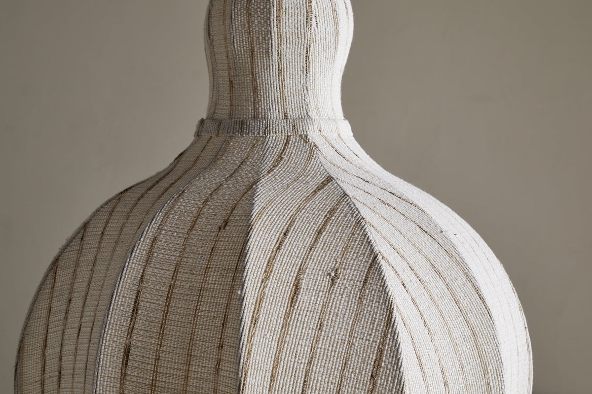 A close-up of the nkuku Kela Fabric Pendant in Off White, featuring a sculptural bulbous base and narrow neck with tightly arranged white beads and thin brown stripes. The handcrafted texture adds a calming ambience against a plain backdrop.