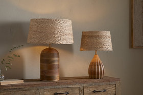 Two nkuku table lamps with Pisang Natural Lampshades sit on a rustic dresser, adding organic texture and warm light. The taller has a rounded dark brown base, while the shorter features a lighter, teardrop-shaped wooden base.