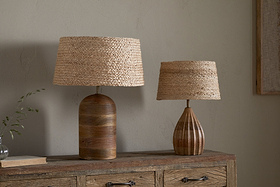 Two table lamps with nkuku Pisang Natural Lampshades sit on a rustic dresser. The left lamp has a rounded, ridged base; the right is smaller with a striped, vase-shaped base. A sprig in a glass and a closed book rest nearby.