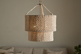 The Thimba Woven Jute Chandelier by nkuku, featuring a double-layered natural jute design, hangs above a neutral couch with pillows, casting a soft glow and rustic texture from matching woven cords.