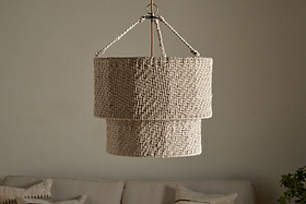 The Thimba Woven Jute Chandelier by nkuku hangs from the ceiling against a plain, light wall. Below, a neutral sofa with textured pillows adds rustic coziness and minimalist charm to the room.