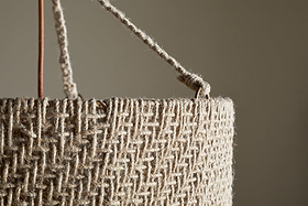 Close-up of the Thimba Woven Jute Chandelier by nkuku, showcasing its rustic texture, zig-zag pattern, and thick rope handle threaded through a metal ring. Set against a neutral beige background with gentle natural light.