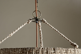 Close-up of the Thimba Woven Jute Chandelier - Natural by nkuku, featuring a rustic natural fiber shade and three braided ropes connecting to a metal ring, suspended from a cord against a plain beige background.
