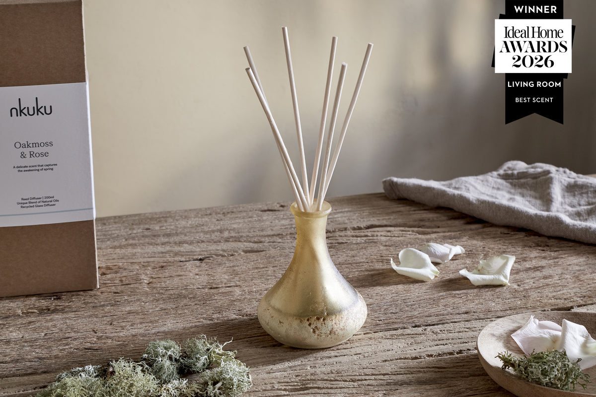 The Oakmoss & Rose Scented Reed Diffuser - Yellow by nkuku, featuring white reeds in hand-blown glass, sits on rustic wood among moss, rose petals, a beige cloth, and its Ideal Home Awards 2026 Best Scent badge.