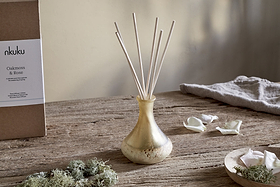An nkuku Oakmoss & Rose Scented Reed Diffuser in yellow sits on a rustic wooden table, its elegant gold vase holding slender reeds that gently diffuse a delicate floral aroma into the air. Surrounding the diffuser are white flower petals arranged on a plate, patches of green moss, a soft grey cloth, and in the background, a branded cardboard box labeled “nkuku Oakmoss & Rose.” The scene is softly illuminated by natural light.