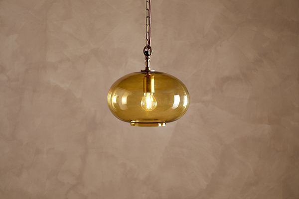 The Otoro Recycled Glass Pendant - Honey - Small Round by nkuku features a single, amber-tinted recycled glass shade paired with elegant brass fittings. Suspended from a chain against a beige, textured wall, the exposed bulb inside casts a warm glow, bringing a cozy, minimalist atmosphere to the space.